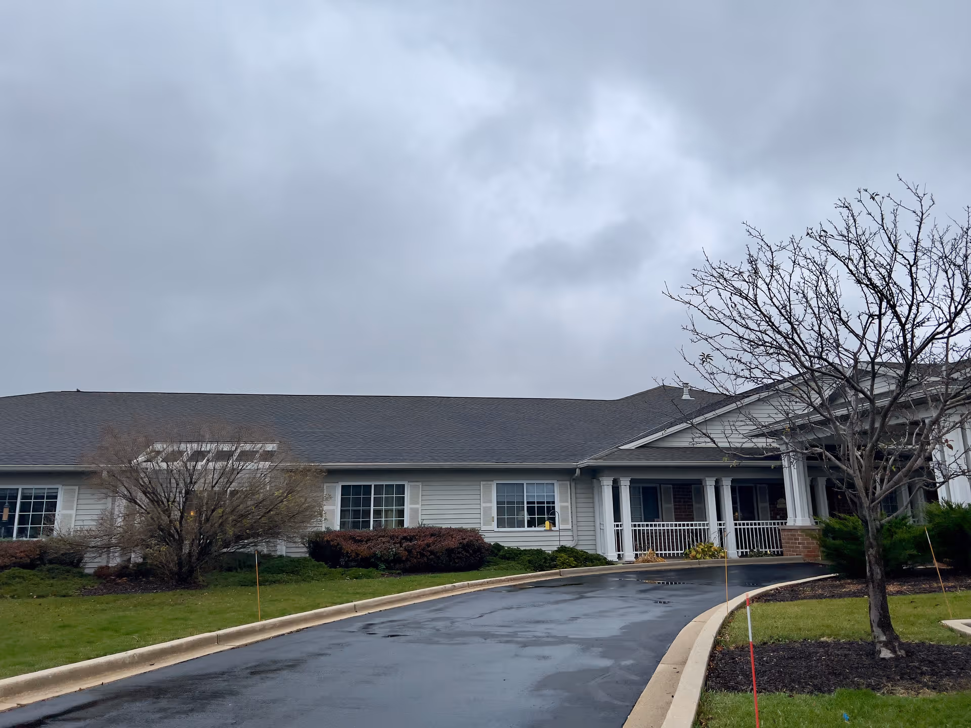 Exterior view of Brenwood Park Assisted Living facility on a cloudy day, showing a single-story building with white siding, multiple windows, a covered entrance with white columns, a curved driveway, and some leafless trees and shrubs in the landscaped front yard.
