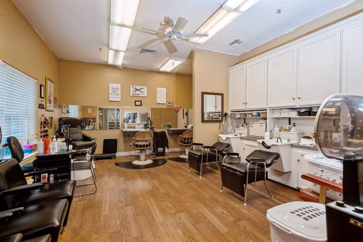 Interior view of a salon area in a senior living facility with multiple black salon chairs, hair washing stations, mirrors on the wall, and shelves with various hair care products. The room has wooden flooring, beige walls, and fluorescent ceiling lights.