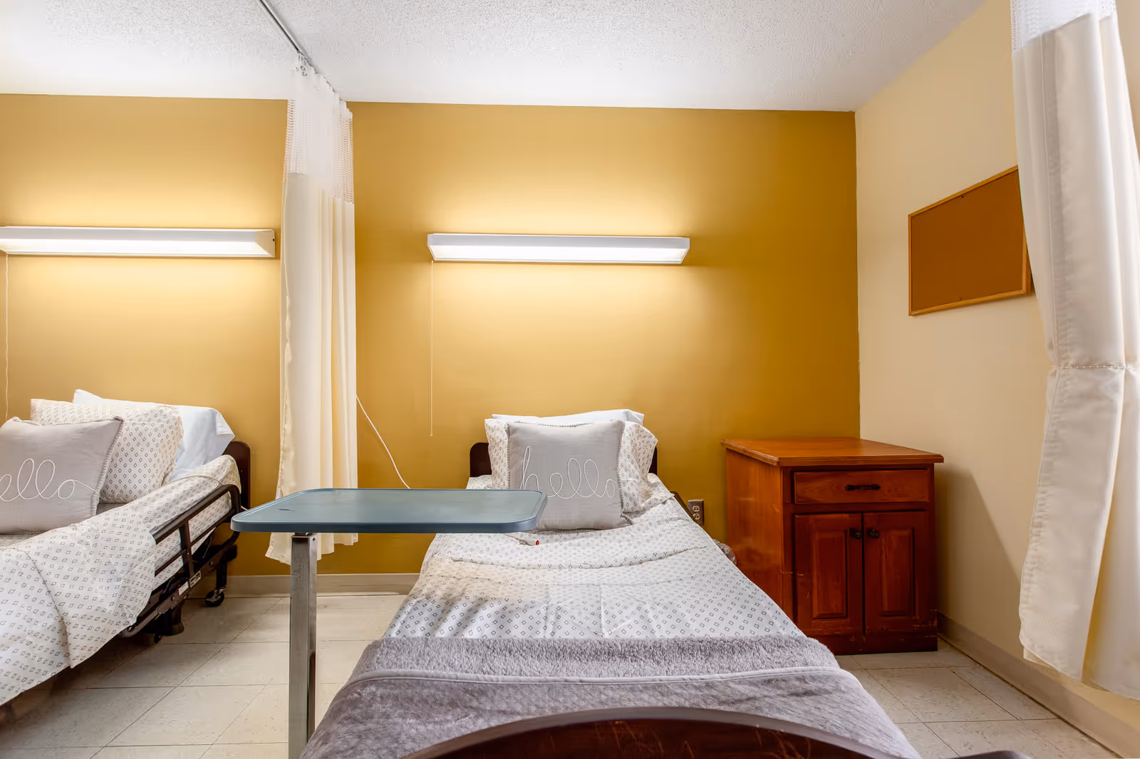 A nursing facility room with two hospital-style beds separated by a curtain. Each bed has white patterned bedding and a gray pillow with the word 'hello' embroidered on it. There is a wooden nightstand next to one bed and a wall-mounted light above each bed. The walls are painted yellow and beige.