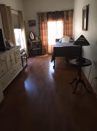 A small bedroom with wooden flooring, a single bed with a pillow and blanket near a window with patterned curtains. There is a wooden side table with a lamp on the right side of the bed, a dresser with a mirror on the left side of the room, and a small vanity table with a round mirror near the window.