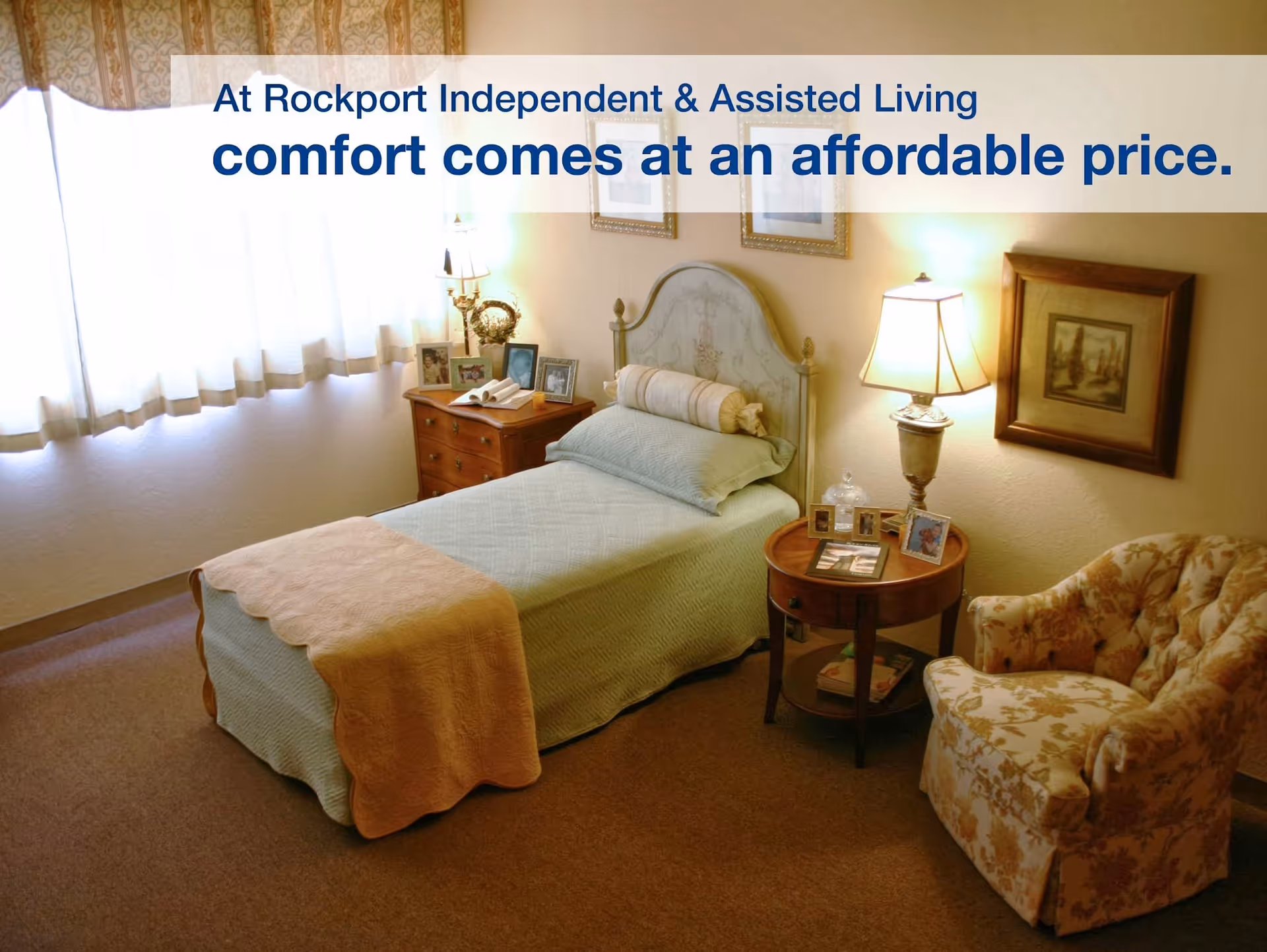 A cozy bedroom in Rockport Retirement Community featuring a single bed with a light green quilt and a peach blanket at the foot. The room has a wooden nightstand with framed photos and a lamp, a floral upholstered armchair, and a window with sheer curtains letting in natural light. There are framed pictures on the walls and a warm, inviting atmosphere.