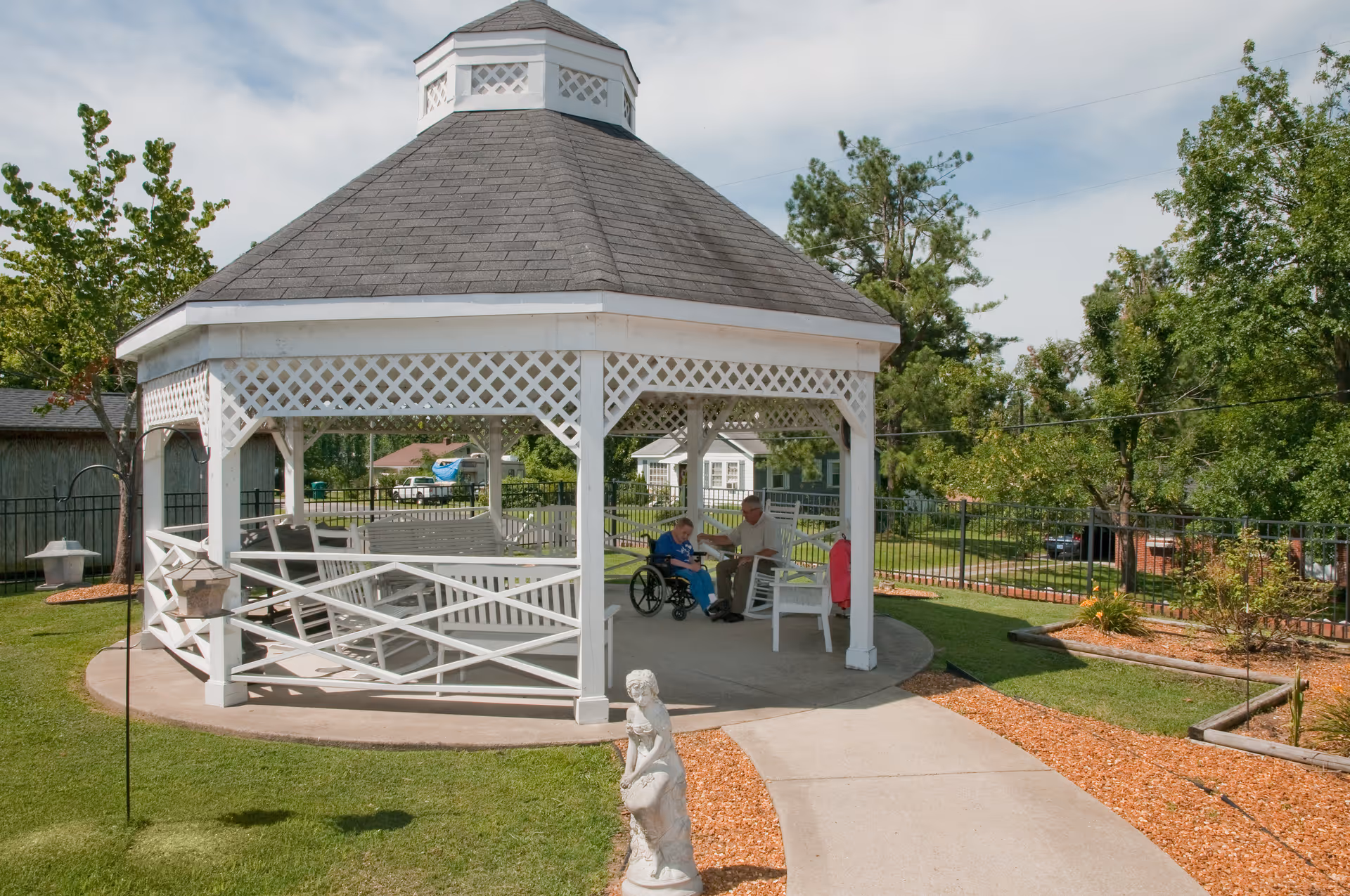 A white gazebo with a shingled roof in a garden area. Inside the gazebo, an elderly person in a wheelchair is accompanied by another person sitting on a chair. The surrounding area has green grass, trees, and a paved walkway leading to the gazebo.