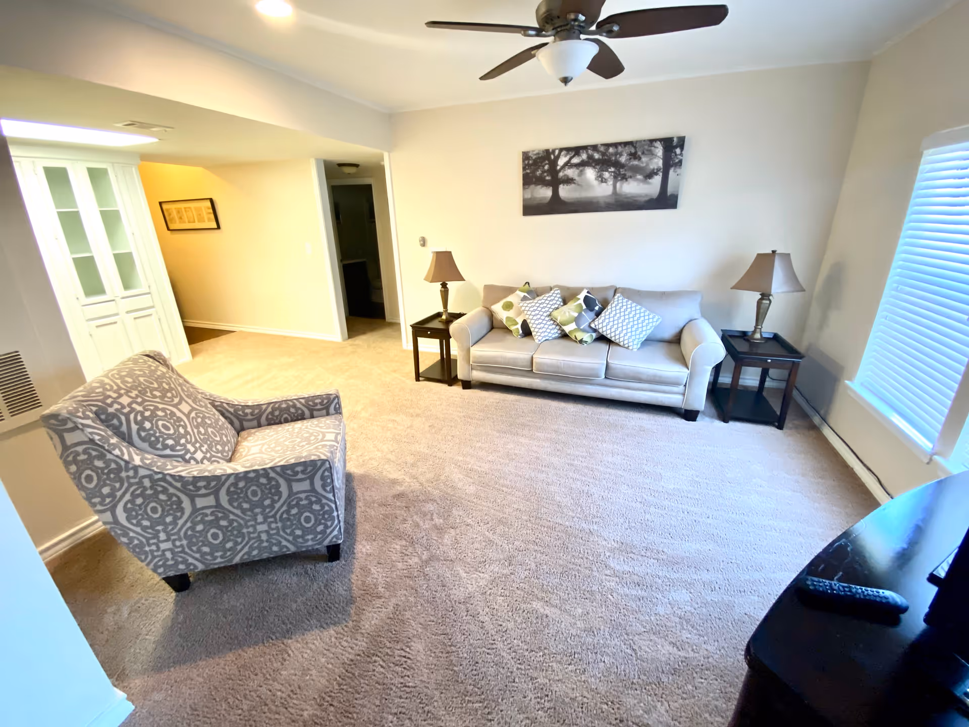 A cozy living room with beige carpet, a light gray sofa adorned with patterned throw pillows, a patterned armchair, two side tables each with a lamp, a ceiling fan, a black and white framed picture of trees on the wall, and a window with blinds letting in natural light.