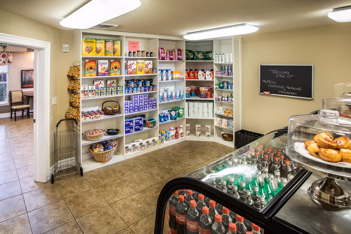 Interior view of a small convenience store area inside a senior living facility named Preston Pointe. Shelves are stocked with various grocery items including cereals, snacks, cleaning supplies, and toiletries. A refrigerated display case in the foreground contains bottled sodas, and a glass display on top holds muffins. A chalkboard on the wall lists the date and muffin flavor. The floor is tiled and there is an open doorway leading to another room with chairs and a table.