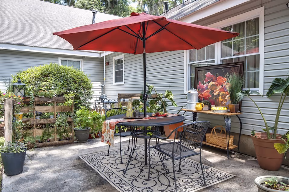 A cozy patio with a red umbrella over a round table and chairs, surrounded by potted plants and decor against the house siding.