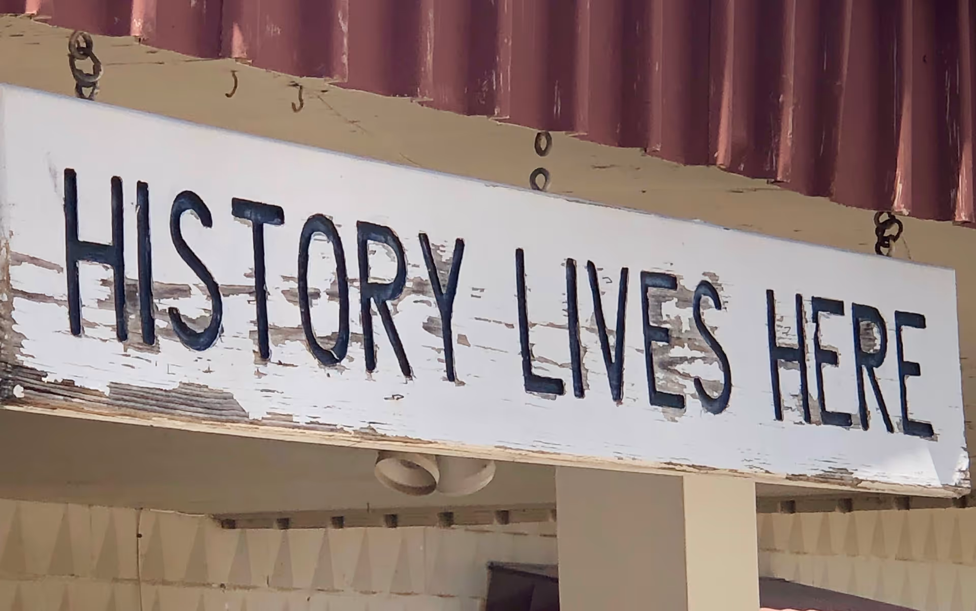 A weathered white wooden sign with black letters that reads 'HISTORY LIVES HERE' hanging under a maroon corrugated roof.
