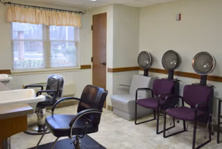 Salon area with barber chairs, hooded hair dryers and extra seating near a window in a senior living facility.