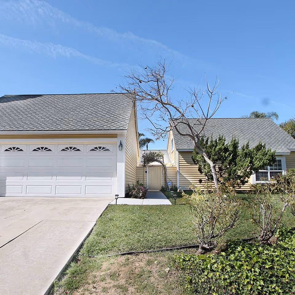 Front exterior of a single-story yellow house with an attached two-car garage, driveway, lawn, and a leafless tree.