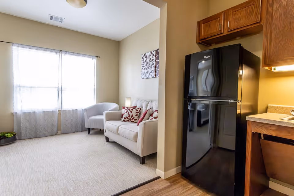 A cozy living area with a beige loveseat adorned with red and white patterned pillows, a white armchair, and a large window with sheer curtains letting in natural light. Adjacent to the living area is a kitchen space featuring a black Whirlpool refrigerator and wooden cabinets.
