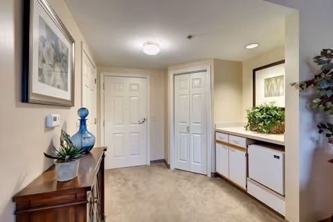 Interior view of a hallway area in a senior living facility featuring a wooden cabinet with decorative items on the left, a white door and closet with double doors straight ahead, and a small kitchenette area with cabinets, a countertop, a dishwasher, and a plant on the right. The walls are light beige and there is a framed picture hanging above the cabinet.