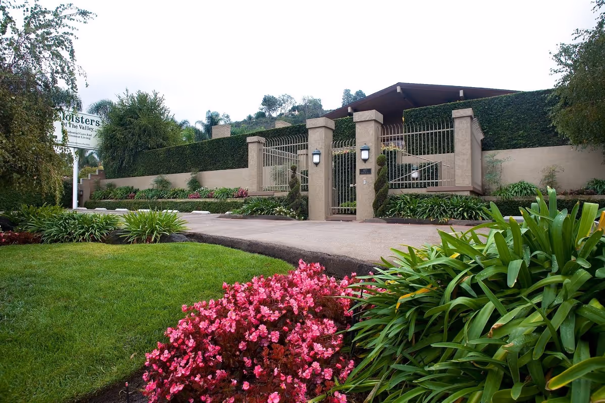 Exterior view of Cloisters Of The Valley facility showing a gated entrance with beige pillars and metal fencing. The entrance is surrounded by well-maintained landscaping including green bushes, pink flowers, and a manicured lawn. A sign with the facility name is visible on the left side.