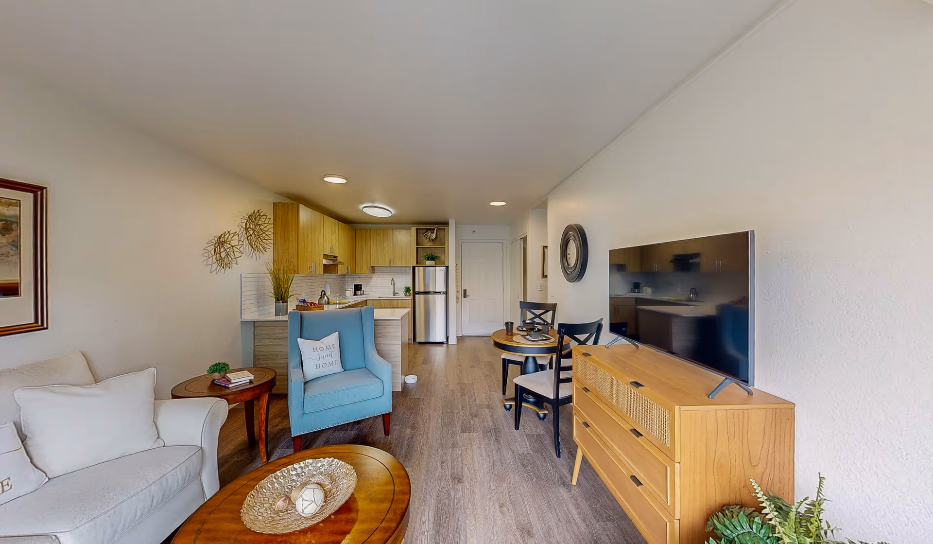 Open-plan living room and kitchenette with a sofa, blue armchair, round coffee table, TV on a wooden dresser, small dining table, and stainless steel refrigerator.