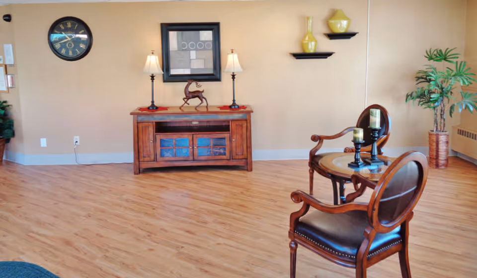 A cozy interior room with wooden flooring featuring a small wooden table with two chairs. On the table are two candle holders with candles. Against the wall is a wooden cabinet with two lamps and a decorative deer figurine. Above the cabinet hangs a framed abstract artwork, and a clock is mounted on the wall to the left. There are two small shelves with yellow vases on the wall and a potted plant in the corner.