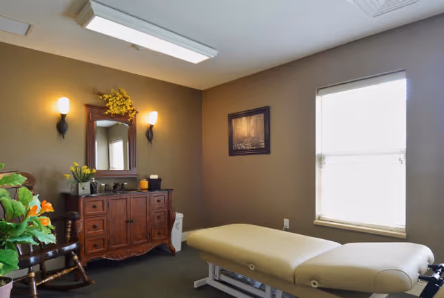 A therapy or massage room with a beige massage table, a wooden cabinet with a mirror and two wall sconces above it, a rocking chair with a cushion, a window with blinds, and a framed picture on the wall. There are also some decorative plants and flowers in the room.