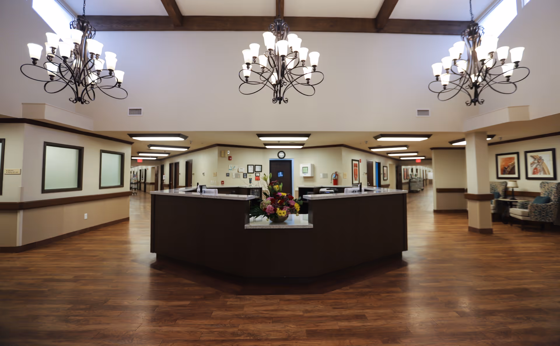 Interior view of a nursing and rehabilitation center reception area with a large dark wood reception desk in the center, wooden flooring, beige walls, decorative framed artwork, and three ornate chandeliers hanging from a high ceiling with exposed wooden beams. Hallways extend from the reception area to the left and right.