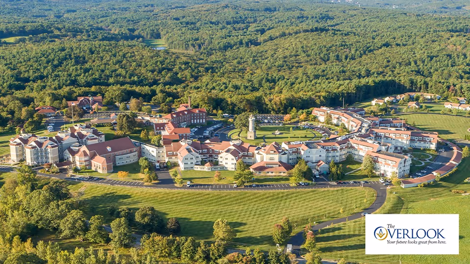 Aerial view of The Overlook senior living facility surrounded by lush greenery and trees. The complex features multiple connected buildings with red roofs, parking areas, and well-maintained lawns. A circular driveway and a central tower or lighthouse structure are visible within the grounds.