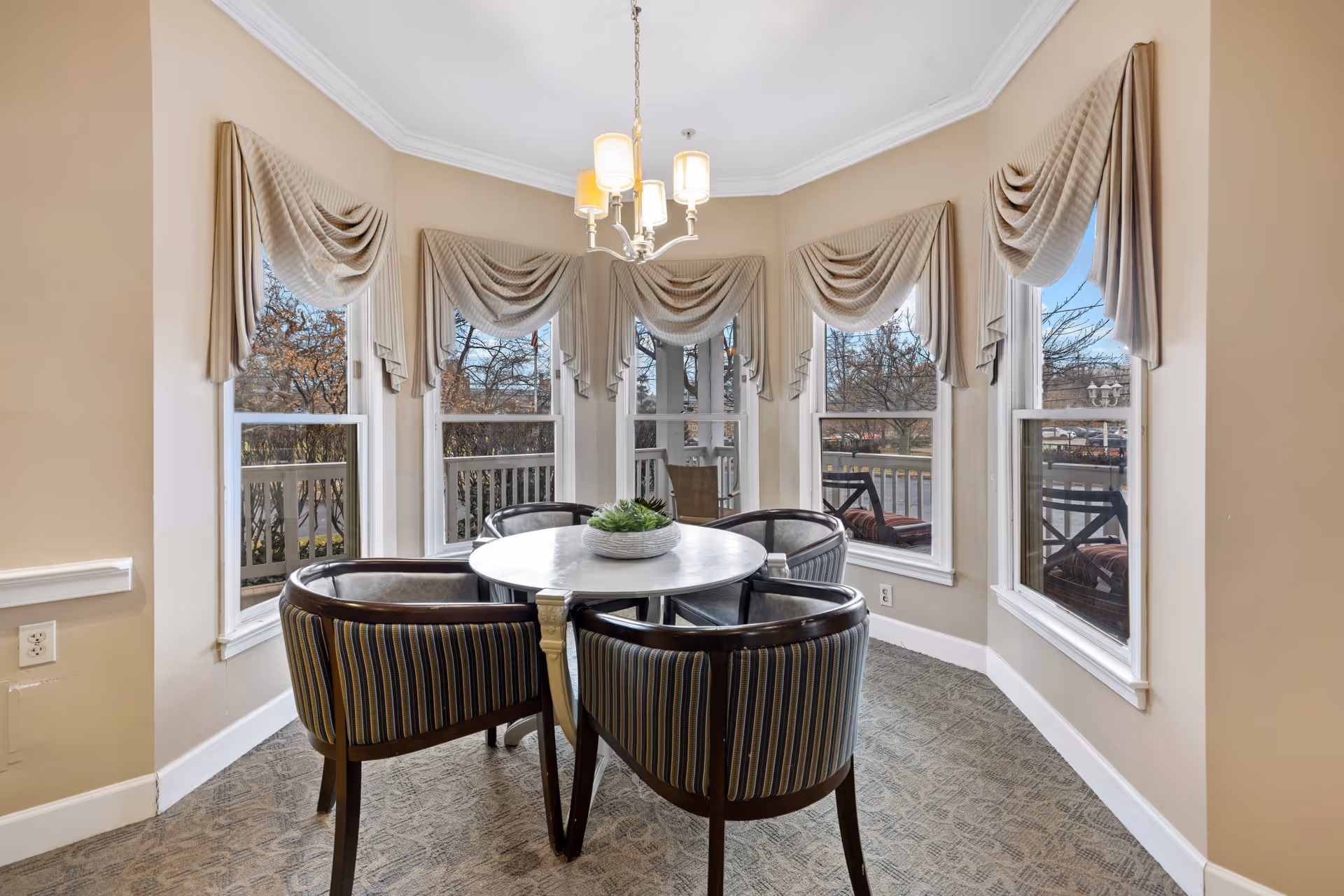 A cozy dining area with a round white table surrounded by four striped upholstered chairs. The space is well-lit by a chandelier hanging from the ceiling and natural light coming through five large windows adorned with beige draped curtains. Outside the windows, trees and a patio area with chairs are visible.