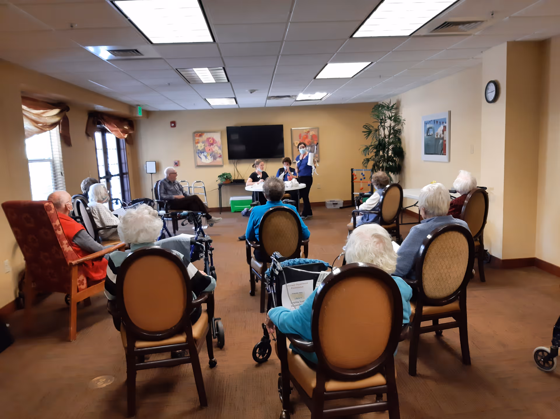 A group of elderly people seated in a semi-circle in a community room, facing two staff members who appear to be leading an activity or presentation. The room has beige walls, a large flat-screen TV mounted on the wall, framed artwork, and a large potted plant. Some residents are using walkers and wheelchairs.