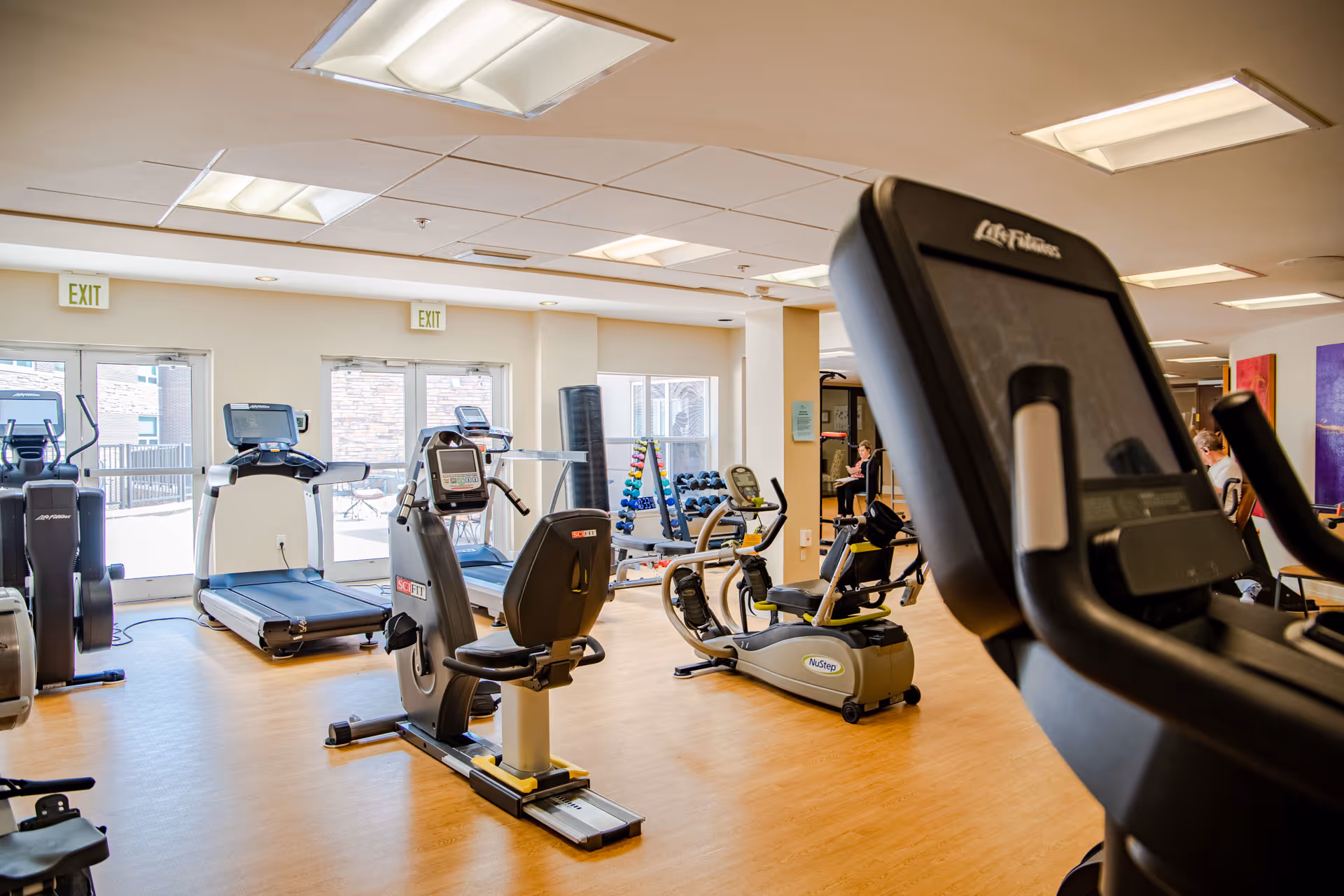 A bright fitness room with exercise equipment including treadmills, stationary bikes, and elliptical machines. The room has large windows and glass doors letting in natural light, light-colored wooden flooring, and a rack of colorful dumbbells in the background.