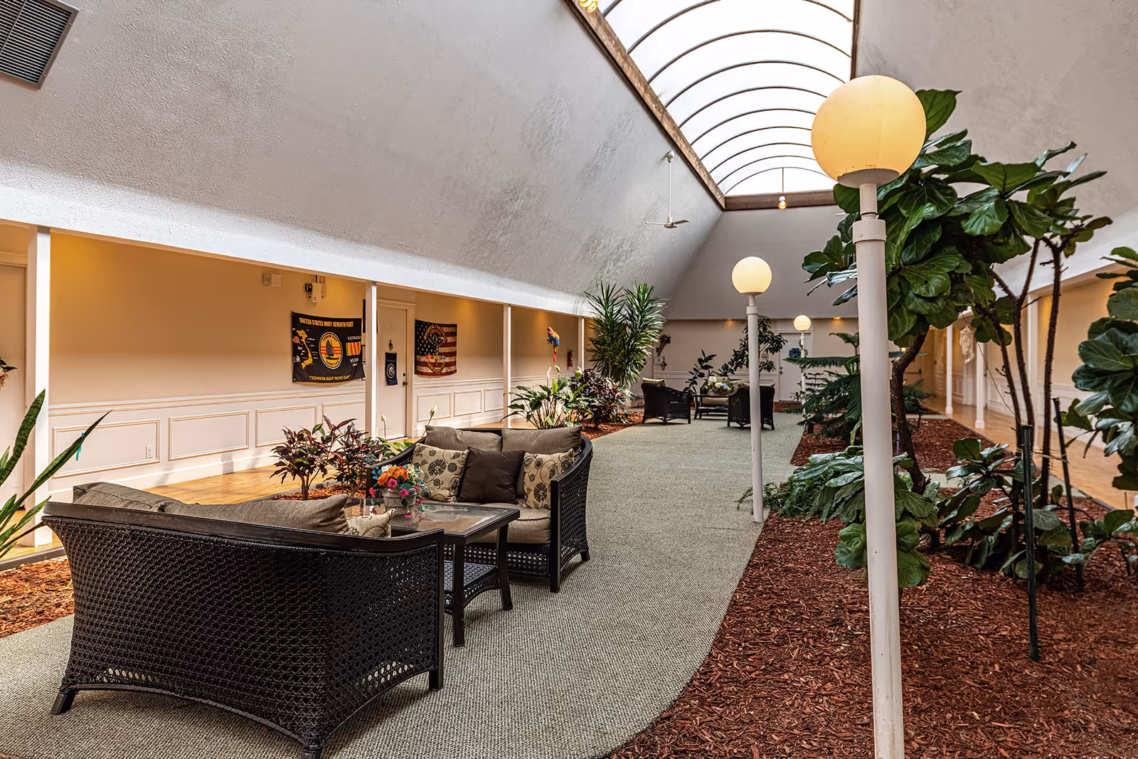 Indoor common area with a high arched skylight ceiling, wicker seating with cushions and a glass-top table, potted plants, and globe-shaped floor lamps along a carpeted walkway. Walls are decorated with flags and plants line the sides of the space.