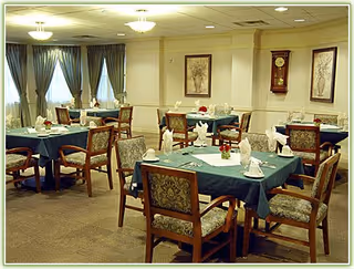 A dining room with several tables covered in dark green tablecloths, each set with white napkins, cups, and small flower arrangements. The room has upholstered wooden chairs, framed artwork on the walls, a grandfather clock, and windows with blue curtains.