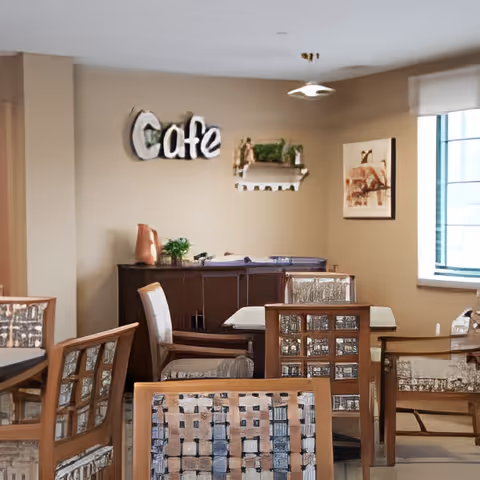 Dining area with wooden tables and upholstered chairs, a 'Cafe' sign on the wall, and a window letting in natural light.