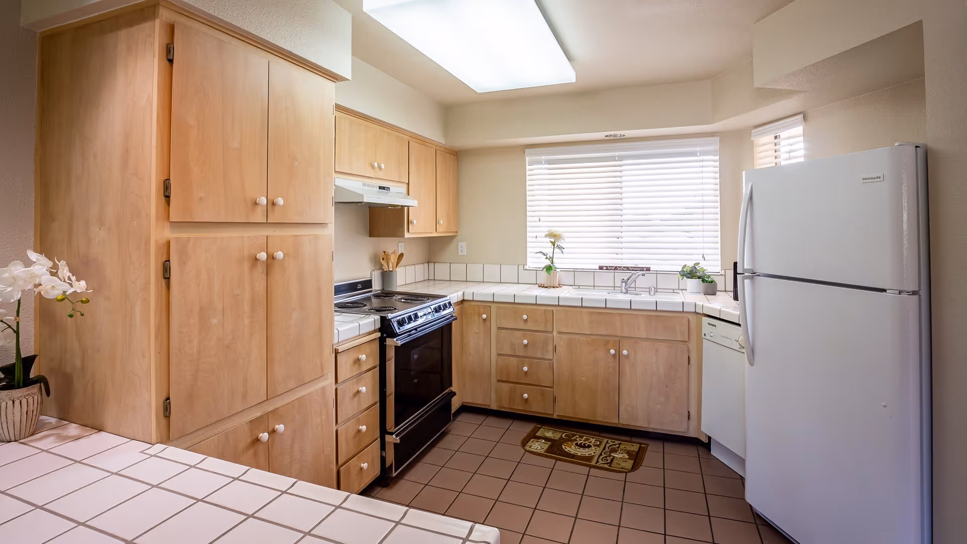 A bright kitchen with light wood cabinets, a white tile countertop, a white refrigerator, a black stove, a dishwasher, and a window with white blinds. There are small plants and kitchen utensils on the countertop.