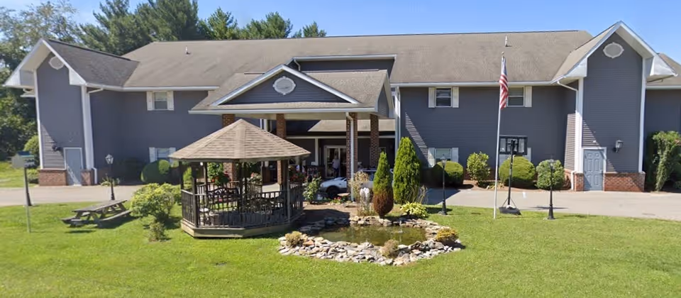 Exterior view of a two-story assisted living facility building with gray siding and a peaked roof. In front of the building is a small landscaped area with a gazebo, a pond surrounded by rocks, shrubs, and an American flag on a flagpole. There is a picnic table on the grass to the left.