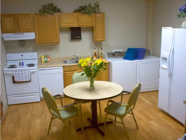 Small communal kitchen with a round table and four chairs, a vase of flowers, stove, sink, refrigerator and a washer and dryer.