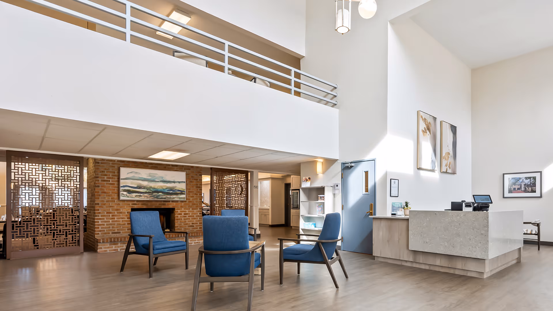 A spacious and bright senior living facility common area with high ceilings, featuring a reception desk on the right, three blue armchairs arranged around a small table in the center, and a brick wall with a fireplace and artwork in the background. Decorative wooden panels partially separate the space.