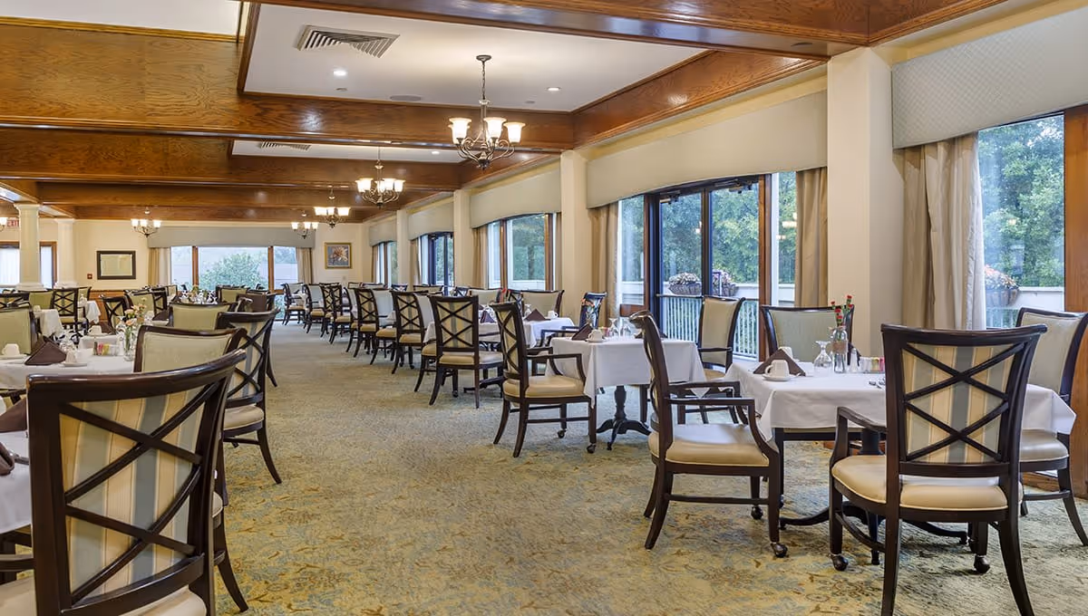 A spacious dining room with multiple tables covered in white tablecloths, each set with napkins, glasses, and silverware. The room features large windows with views of greenery outside, wooden ceiling beams, and chandeliers providing warm lighting. Chairs with cushioned seats and backs are arranged around the tables.