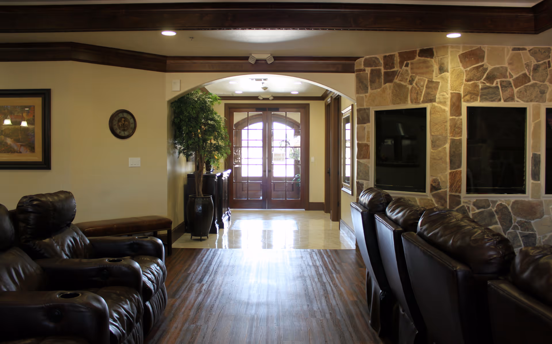 Interior view of a senior living facility lounge area with dark leather recliner chairs arranged in rows facing a stone wall with built-in TV spaces. The room has wooden flooring, a potted plant near an archway, and double glass doors letting in natural light.