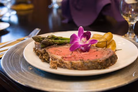 A plated meal featuring a slice of roast beef garnished with a purple orchid flower, accompanied by asparagus and roasted potatoes, served on a white plate with a decorative charger underneath on a dining table.