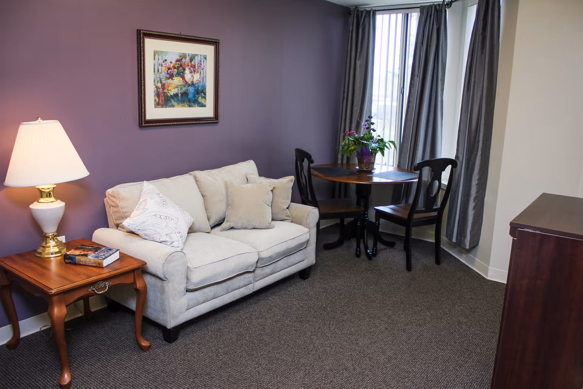 A cozy sitting area in an assisted living facility with a beige loveseat adorned with three cushions, a wooden side table with a lamp and a book, a round wooden table with two chairs, a potted plant on the table, and a framed floral painting on a purple wall.