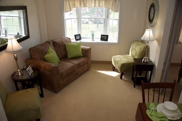 Small living room with a brown loveseat, green accent chairs and pillows, bay window, lamps, and side tables.
