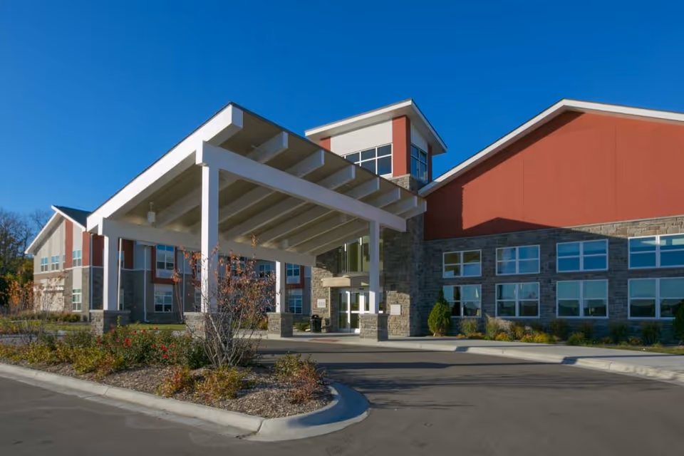 Exterior view of The Healthcare Resort Of Topeka building with a large covered entrance, stone and red siding, multiple windows, and landscaped plants in front under a clear blue sky.