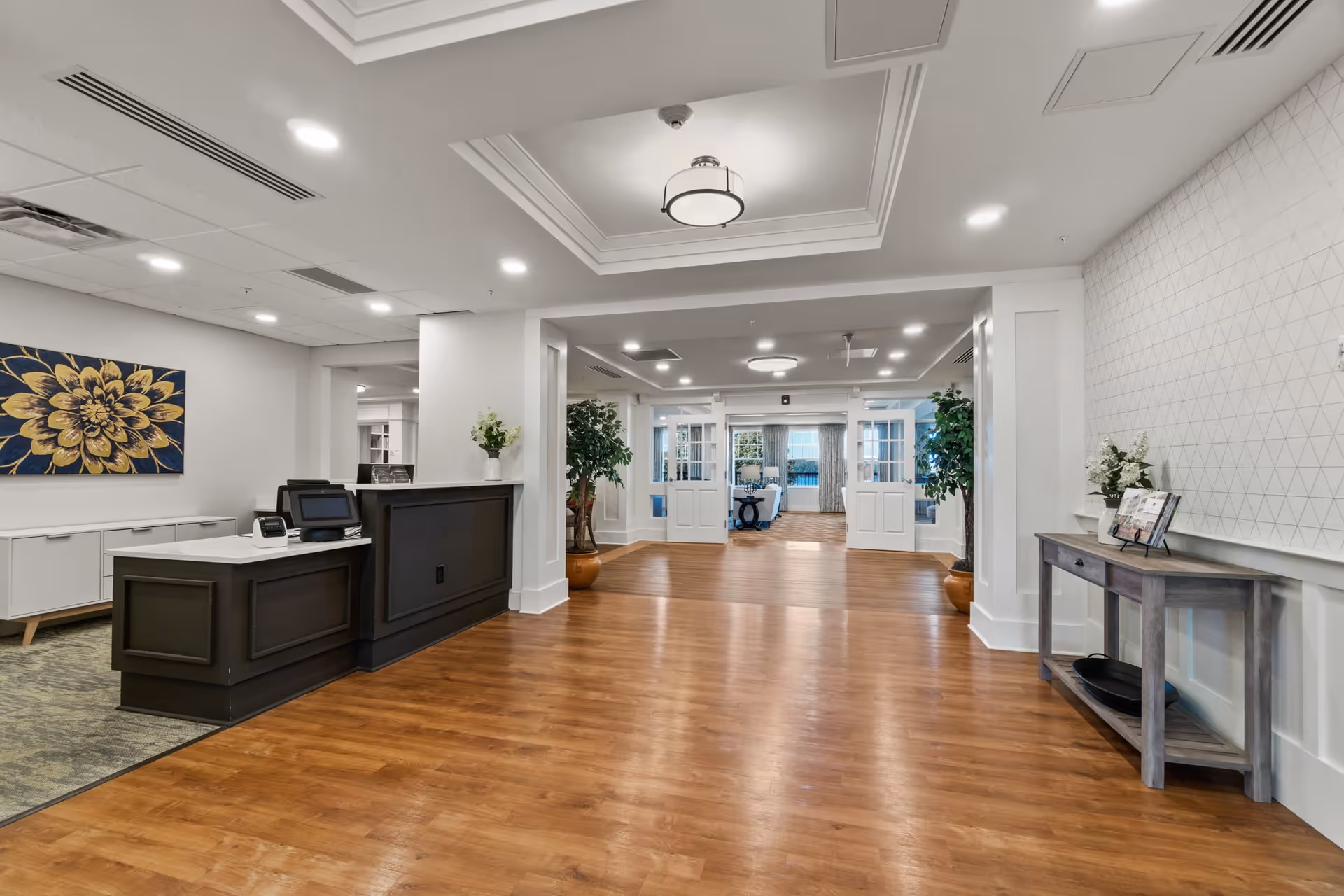 Spacious and well-lit senior living facility interior with wooden flooring, a reception desk on the left, decorative plants, and a hallway leading to a seating area with large windows.