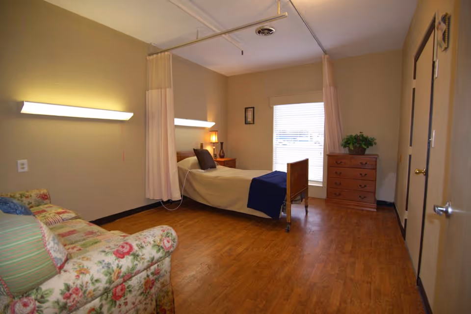 A nursing facility bedroom with a single bed covered in beige bedding and a dark blue blanket at the foot. The bed has a headboard and footboard and is positioned near a window with blinds. There is a wooden dresser with a potted plant on top to the right of the window. A floral-patterned armchair with striped and floral cushions is placed on the left side of the room. The room has wood flooring, beige walls, two wall-mounted lights, and a ceiling-mounted privacy curtain partially drawn around the bed.