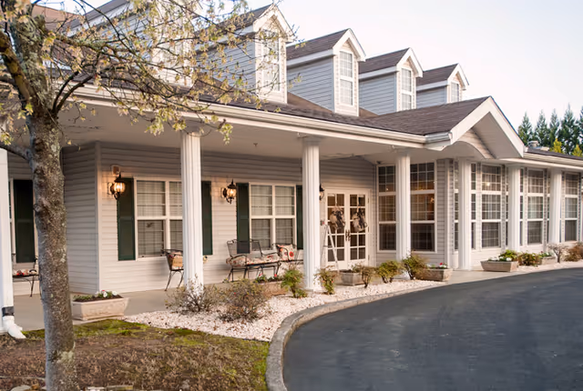 Exterior view of a senior living facility with a covered porch supported by white columns, several windows with green shutters, benches on the porch, and a curved driveway in front. There are some small plants and a tree near the building.