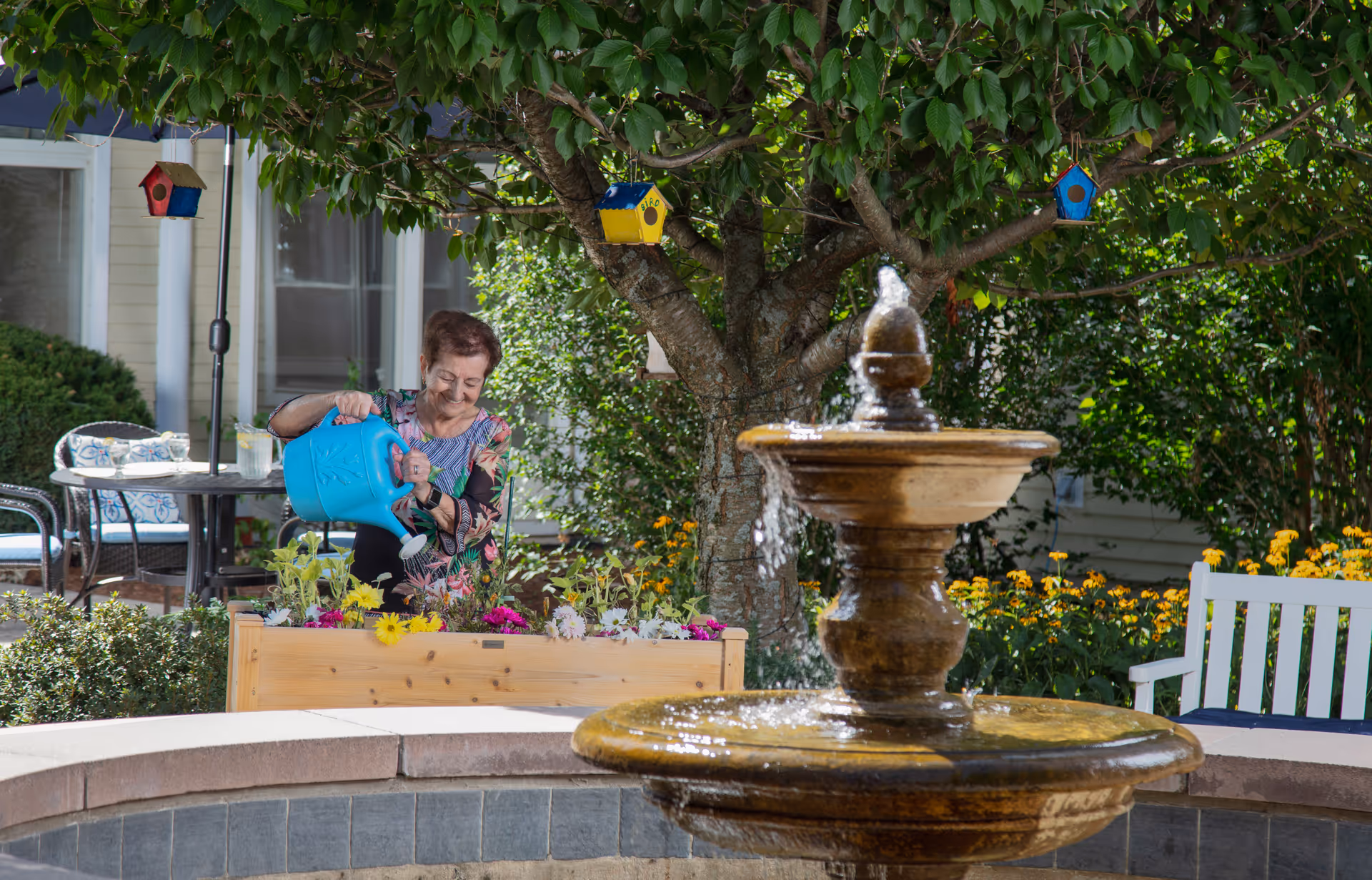 A woman waters flowers in a raised planter beside a tree and a decorative fountain in an outdoor garden courtyard.