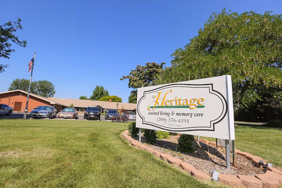 Exterior view of the Heritage Assisted Living sign on a lawn with the facility building, parked cars, and an American flag in the background.