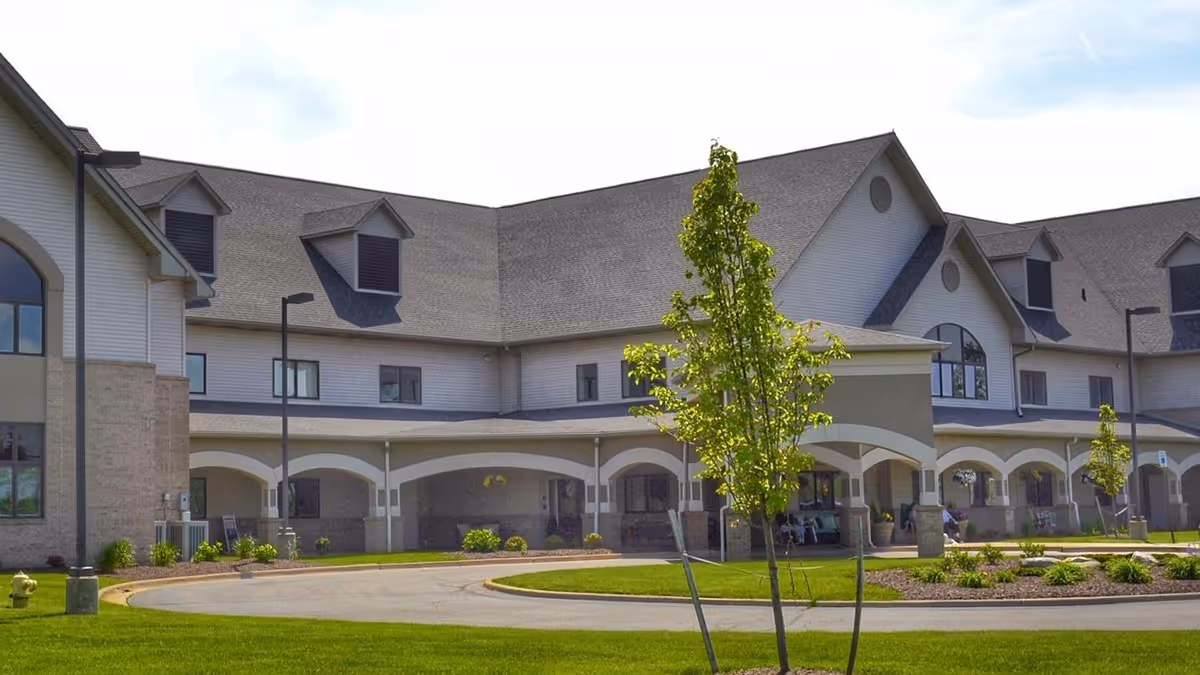 Front exterior of a large multi-story retirement facility with an arched covered entry, driveway, and landscaped lawn.