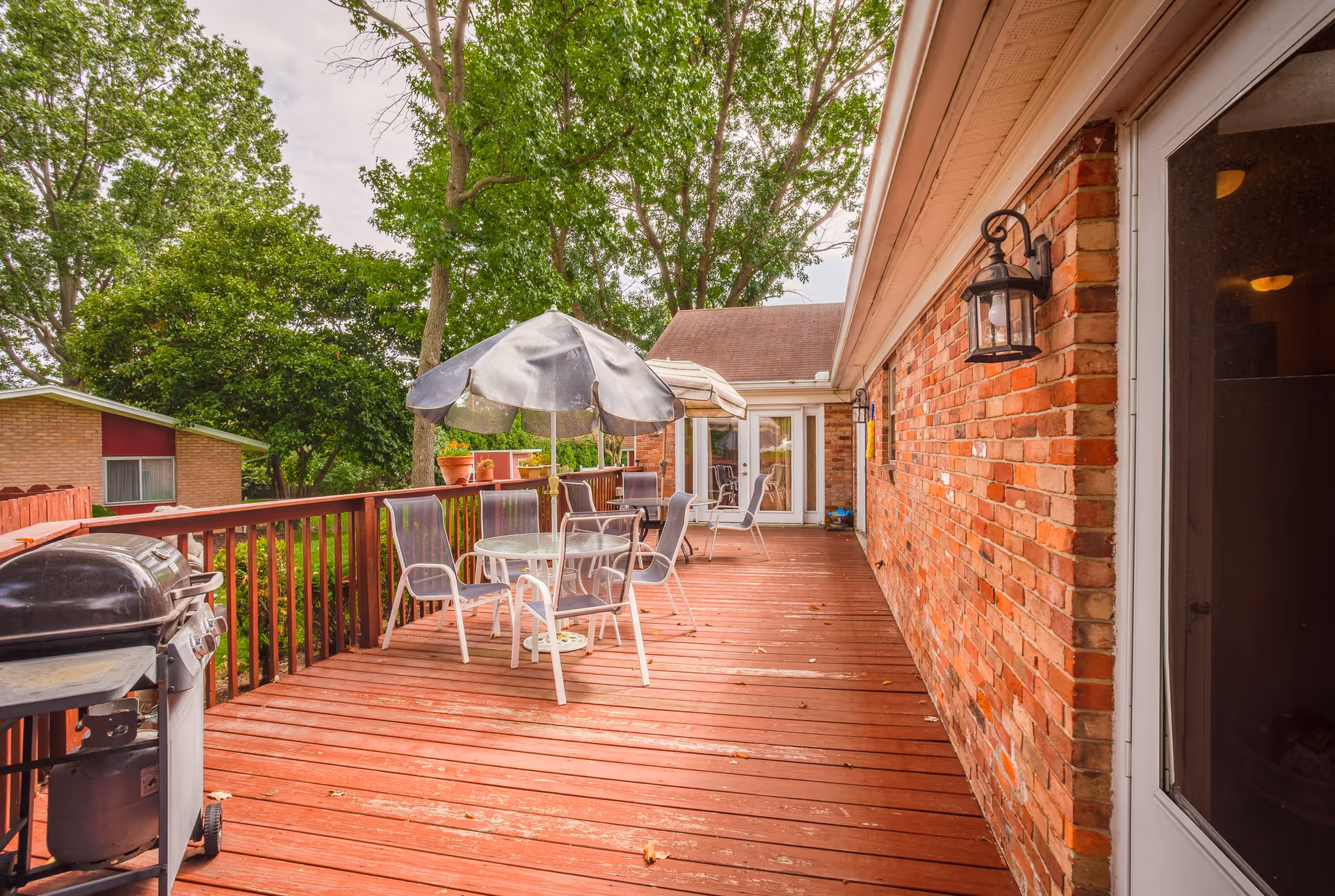 Outdoor wooden deck attached to a brick building with patio furniture including tables, chairs, and umbrellas. There is a grill on the left side and trees and greenery surrounding the area.