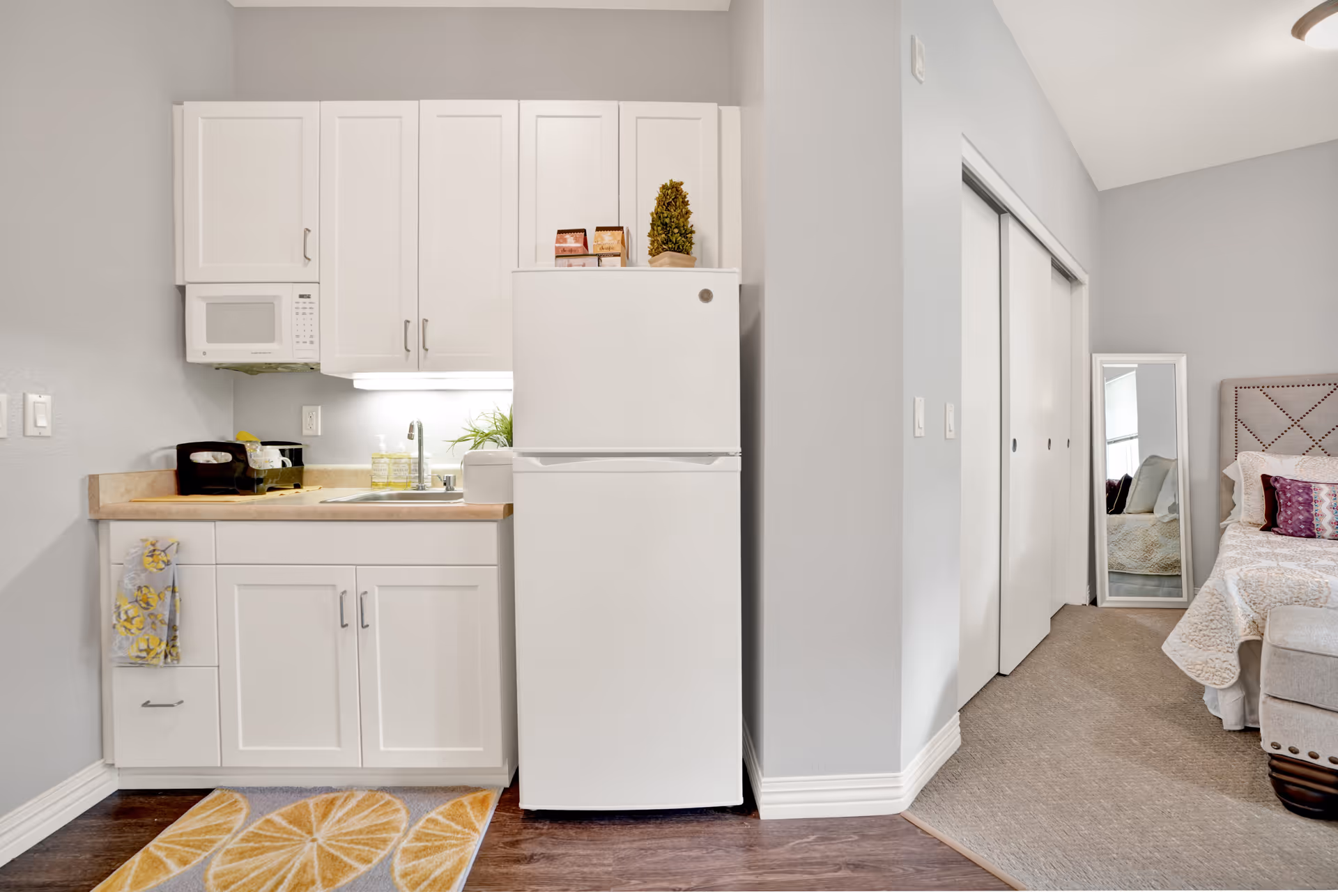 Small studio kitchenette with white cabinets, a refrigerator and sink, adjacent to a visible bedroom area.