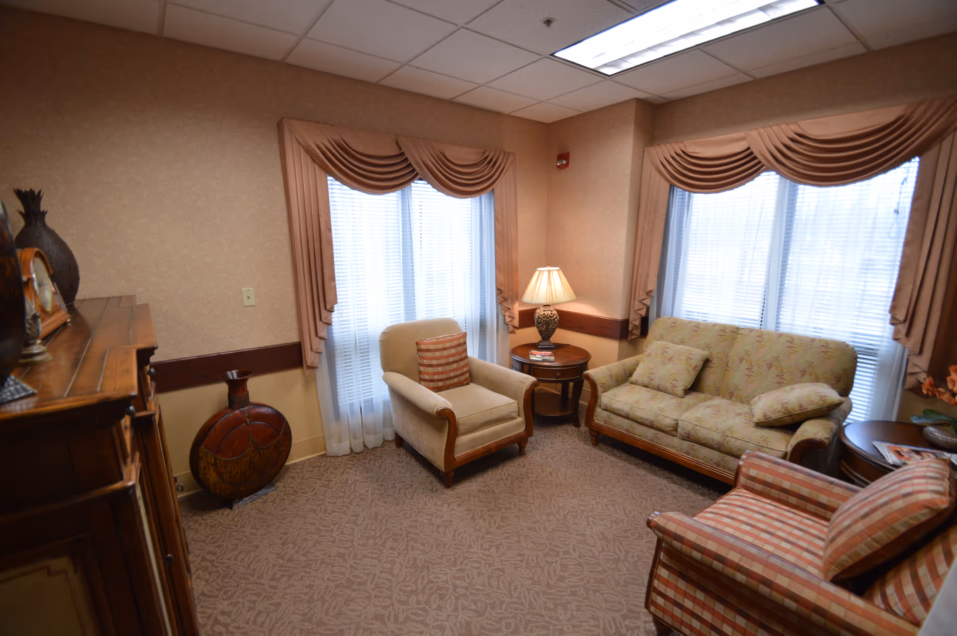 A cozy living room with beige walls and carpet, featuring a floral-patterned sofa with two cushions, a beige armchair with a striped cushion, and a striped armchair. There are two large windows with sheer white curtains and brown drapes. A round wooden side table with a lamp and magazines is placed between the sofa and armchair. A wooden cabinet and decorative vases are visible on the left side of the room.