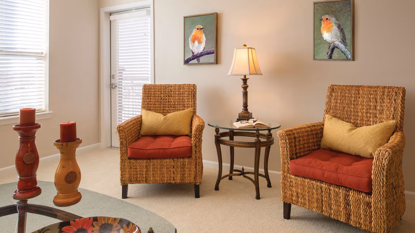 A cozy living room area with two wicker armchairs featuring red cushions and yellow pillows. Between the chairs is a round glass-top side table with a lamp, an open book, and a pair of glasses. On the wall above each chair hangs a framed painting of a small bird. To the left, there are two decorative candle holders with red candles near a window with white blinds and a glass door with blinds.
