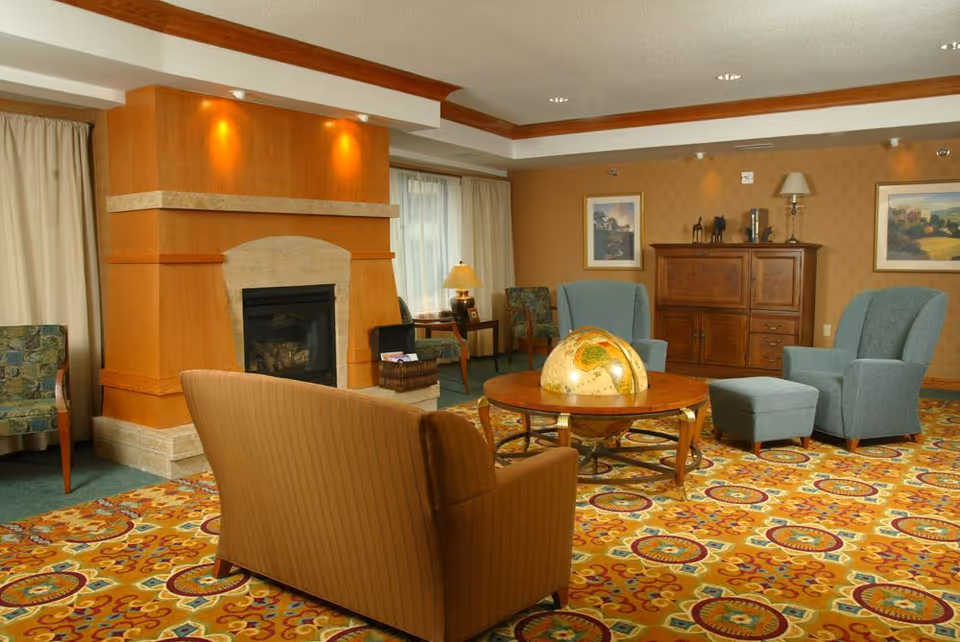 A cozy living room with a patterned carpet, a fireplace with a wooden mantle, a round wooden coffee table with a globe on it, a brown sofa, two blue armchairs with a matching ottoman, a wooden cabinet, framed artwork on the walls, and a table lamp near a window with curtains.
