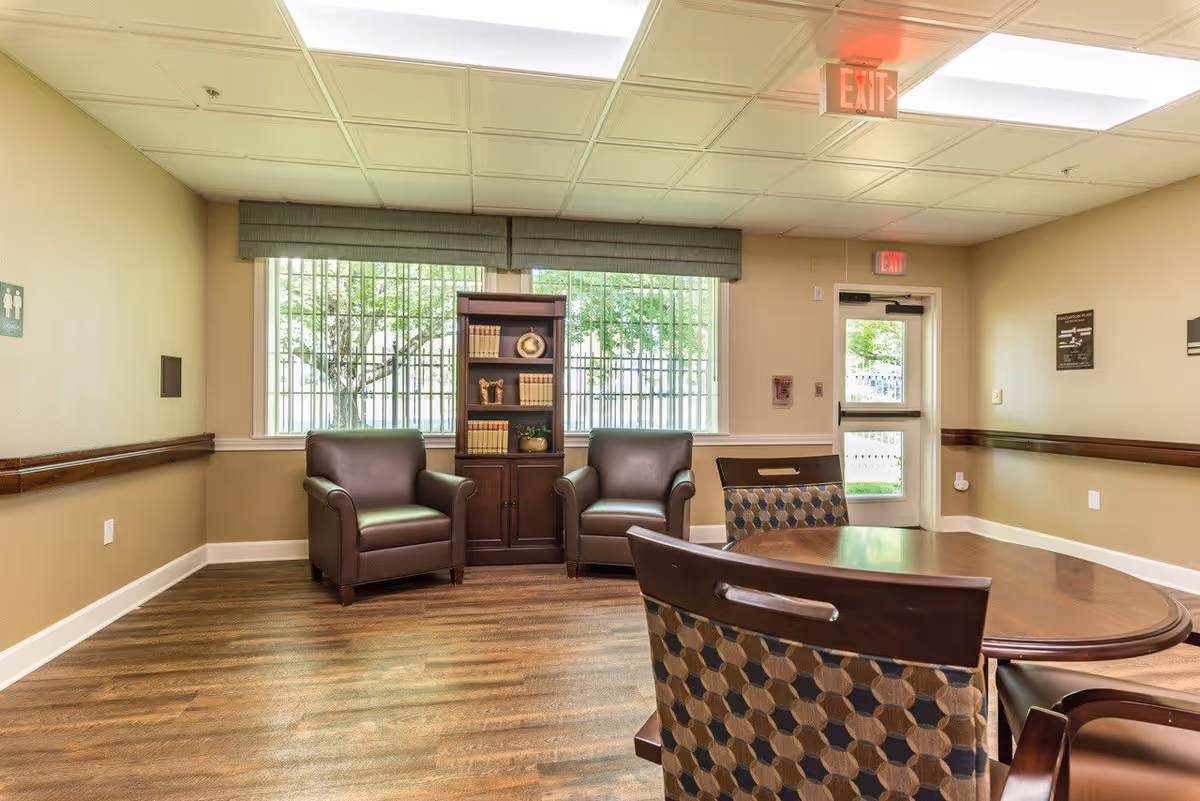 A well-lit interior room with two brown leather armchairs and a wooden bookshelf filled with books and decorative items placed between two large windows. In the foreground, there is a round wooden table with patterned cushioned chairs. The room has wood flooring, beige walls, and an exit door with a red exit sign above it.