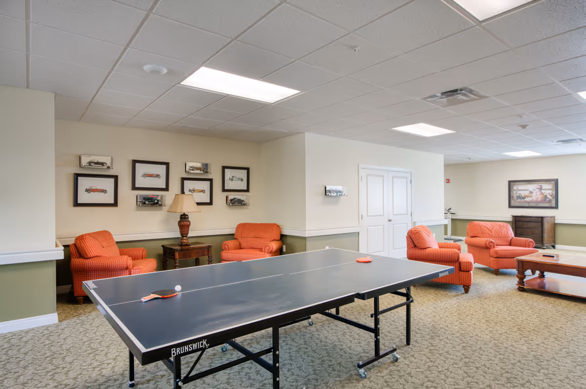 A spacious common room with a ping pong table in the center. The room features four orange upholstered armchairs arranged around wooden side tables and a coffee table. The walls are decorated with framed pictures of vintage cars and a painting. The ceiling has recessed lighting and a drop ceiling design.