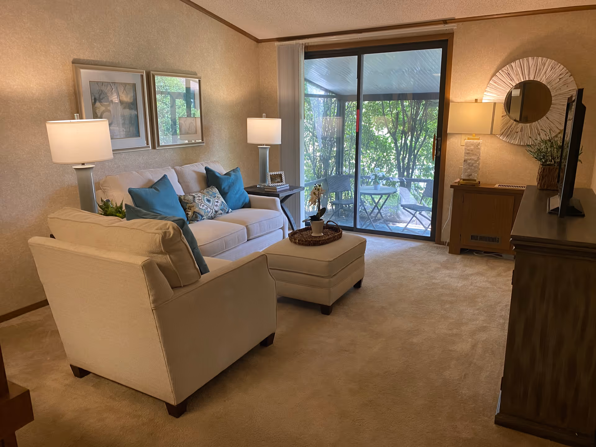 Cozy living room with a beige sofa, armchair, ottoman, lamps, and a sliding glass door leading to a small patio.